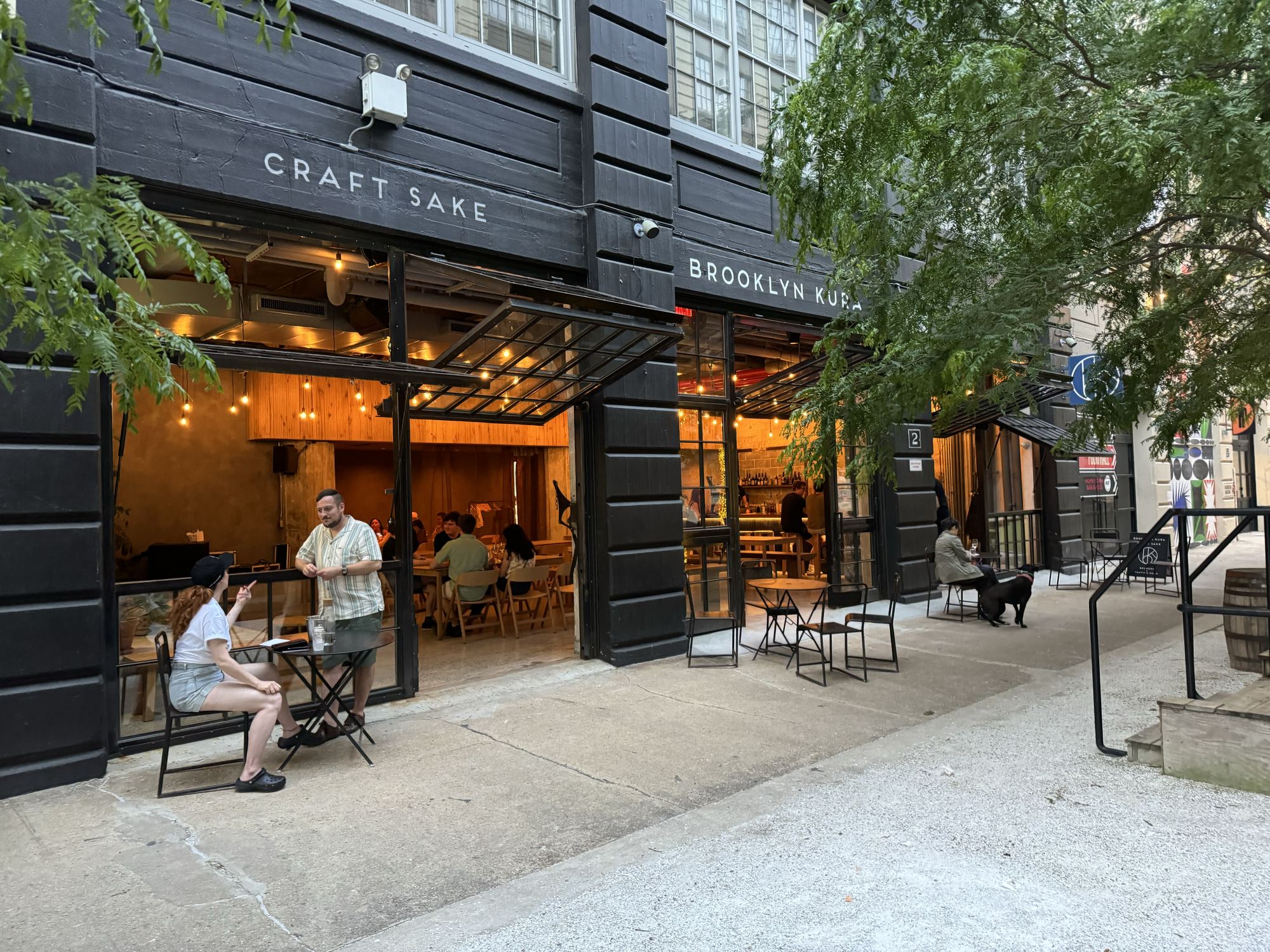 The Brooklyn Kura taproom at dusk, doors open and lights glowing, with guests at sidewalk tables