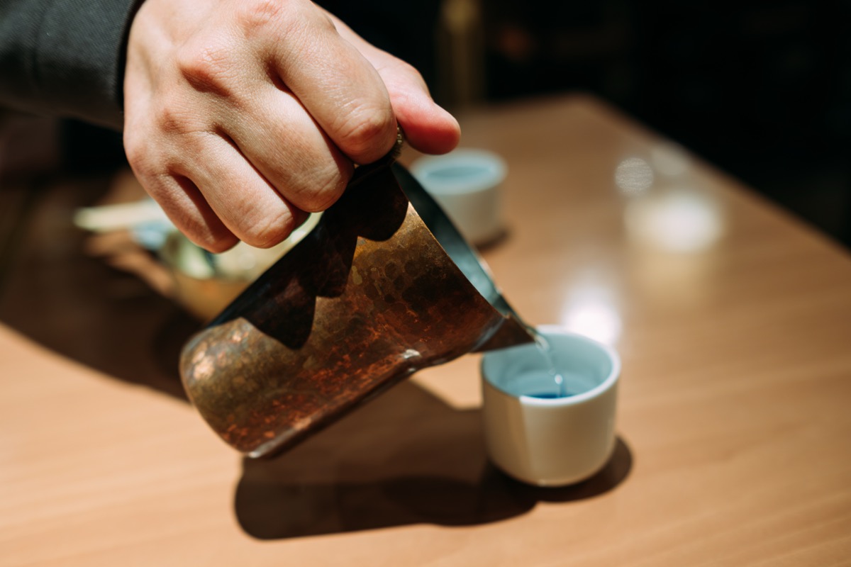 Pouring warm sake from a copper warmer into a tasting cup
