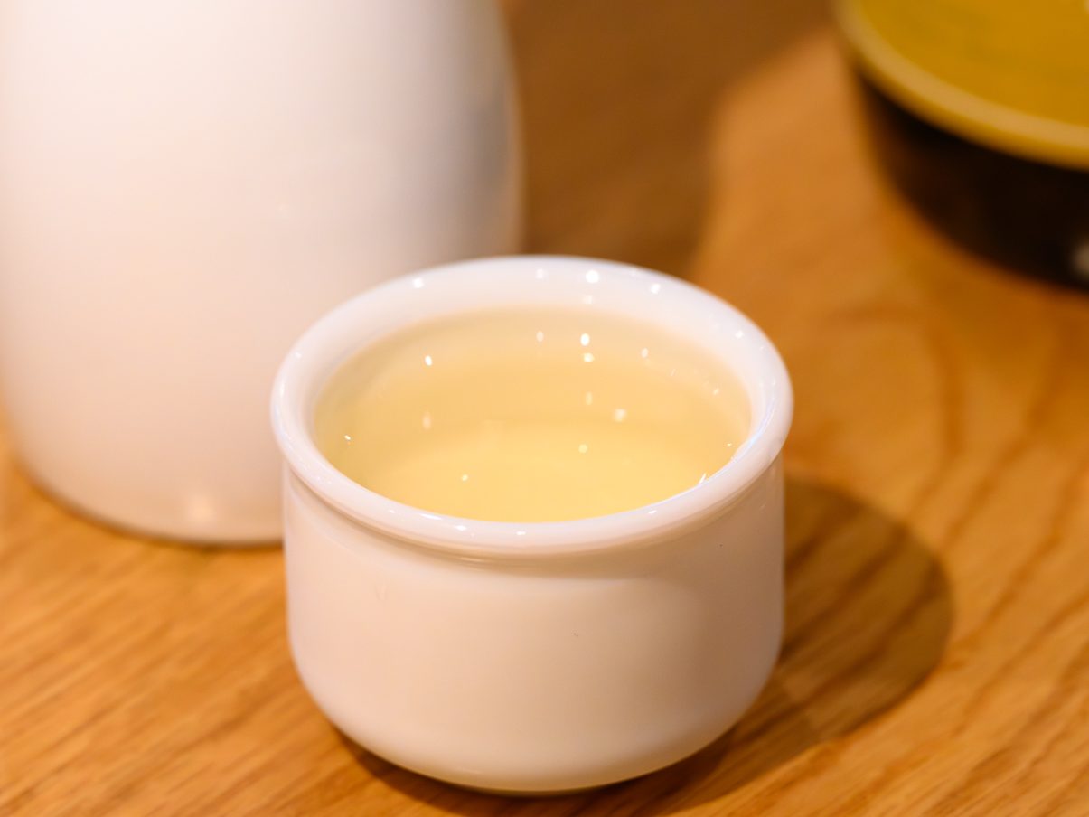 A cup of golden warm sake beside a white tokkuri carafe